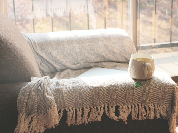 Eine gemütliche Couch mit einer Wolldecke und einer Tasse Tee steht vor einem Fenster mit Blick auf eine verschneite Landschaft.